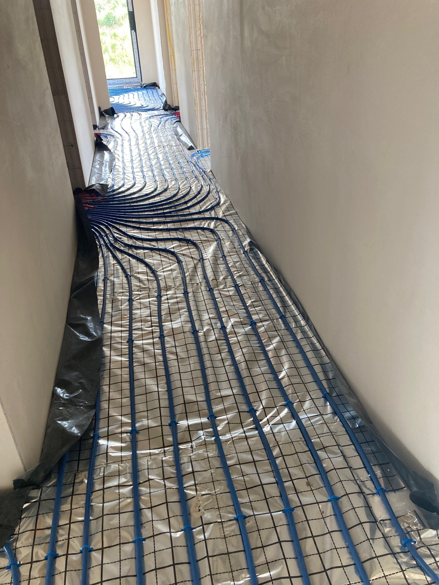 Niebieskie rurki ogrzewania podłogowego ułożone na srebrnej folii izolacyjnej z czarną siatką, widok w korytarzu w trakcie instalacji.