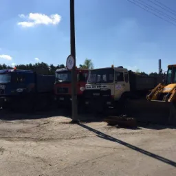 Rząd ciężarówek MAN i IVECO oraz żółta koparko-ładowarka JCB stojące na placu budowy w słoneczny dzień, widoczny słupek z ograniczeniem prędkości do 15 km/h.