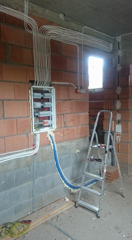 Rozdzielnica elektryczna w stanie surowym, widoczne liczne przewody w peszlach, drabina aluminiowa i narzędzia.