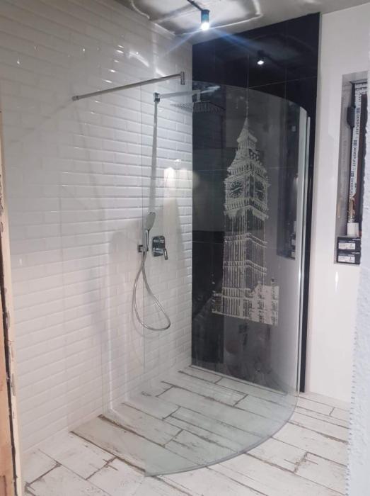 Nowoczesna kabina prysznicowa z zaokrągloną szybą, białe kafelki imitujące cegłę, drewnopodobna podłoga i dekoracyjny panel z motywem Big Bena.