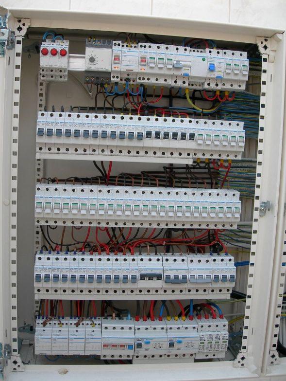 Otwarta, biała skrzynka rozdzielcza z zamontowanymi bezpiecznikami i przewodami elektrycznymi w różnych kolorach, widoczne liczne wyłączniki nadprądowe i różnicowoprądowe.