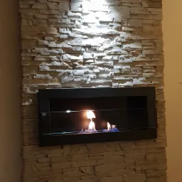 Ściana z dekoracyjnego kamienia z wbudowanym biokominkiem z czarną ramą i widocznym płomieniem, oświetlona od góry punktowym światłem.