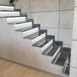 Schody z granitu z podświetleniem LED, ściana wykończona dekoracyjnym tynkiem w odcieniach szarości. Nowoczesne wykończenie wnętrza z dbałością o detale.