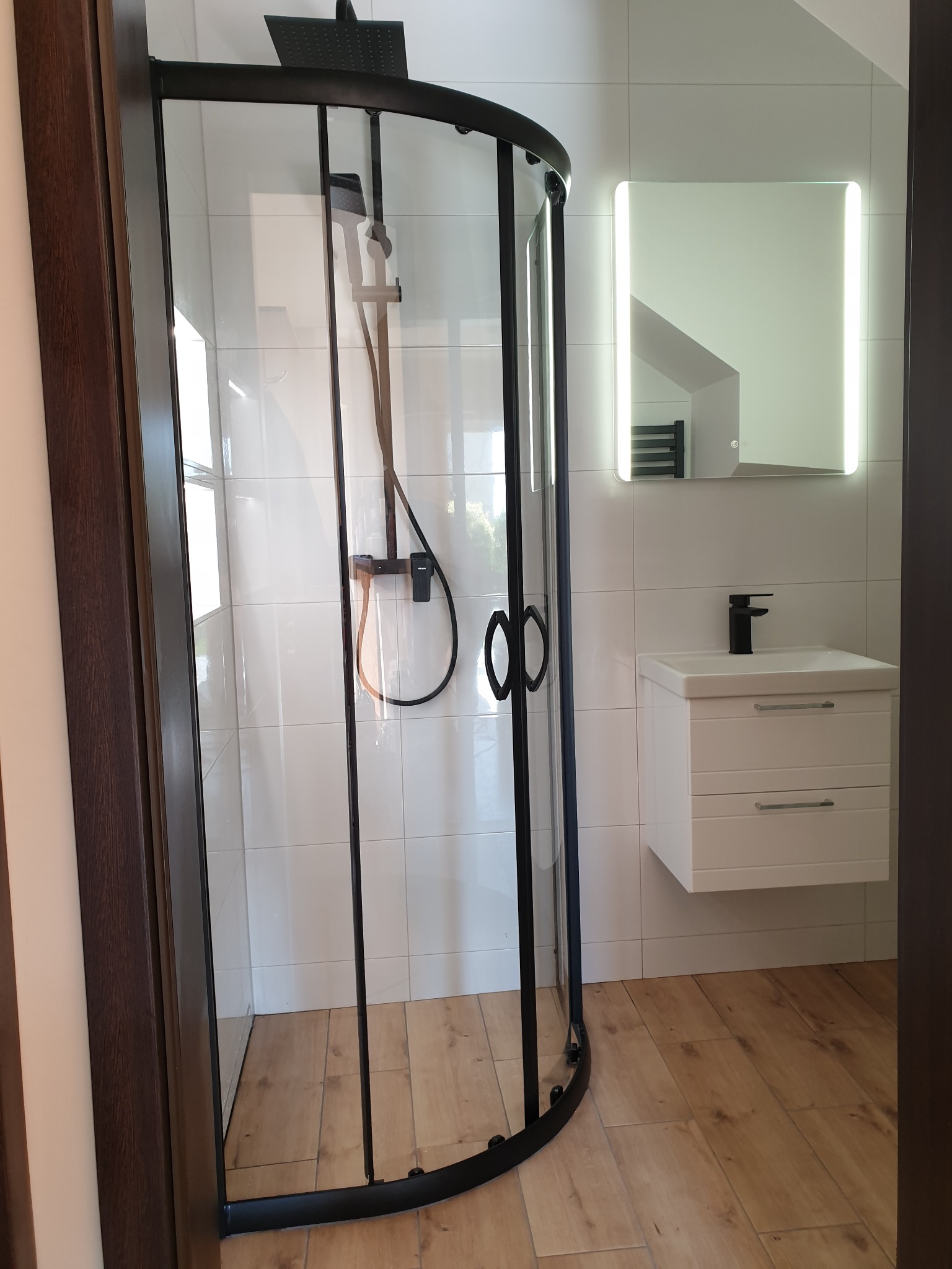 Nowoczesna łazienka z kabiną prysznicową typu walk-in z czarnymi profilami, białą umywalką z szafką i podświetlanym lustrem na białych płytkach, z drewnopodobną podłogą.