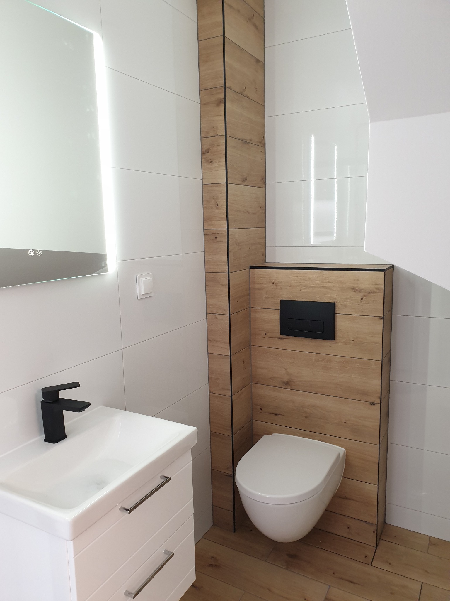 Nowoczesna łazienka z podwieszaną toaletą w obudowie imitującej drewno, białą umywalką z czarną baterią i podświetlanym lustrem na tle białych płytek.