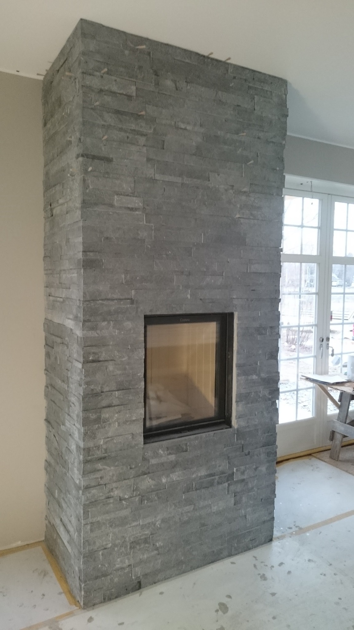 Obudowa kominka wyłożona szarym kamieniem łupkowym, widoczna szyba kominka oraz fragment okna balkonowego w tle.
