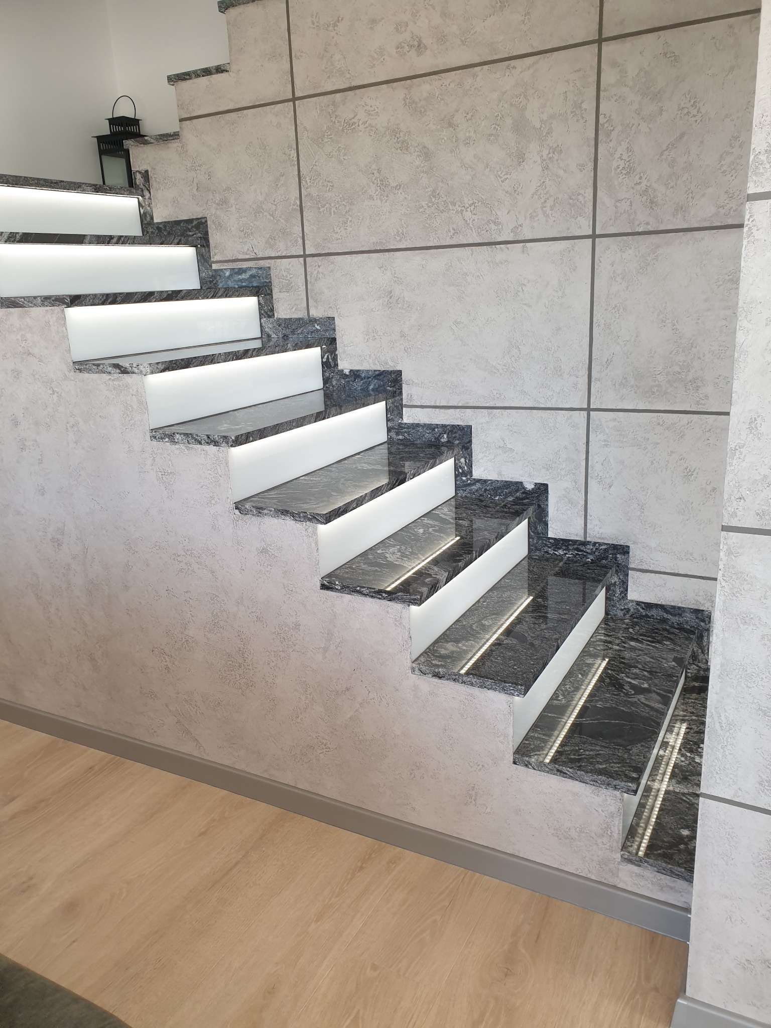 Schody z granitu z podświetleniem LED, ściana wykończona dekoracyjnym tynkiem w odcieniach szarości. Nowoczesne wykończenie wnętrza z dbałością o detale.