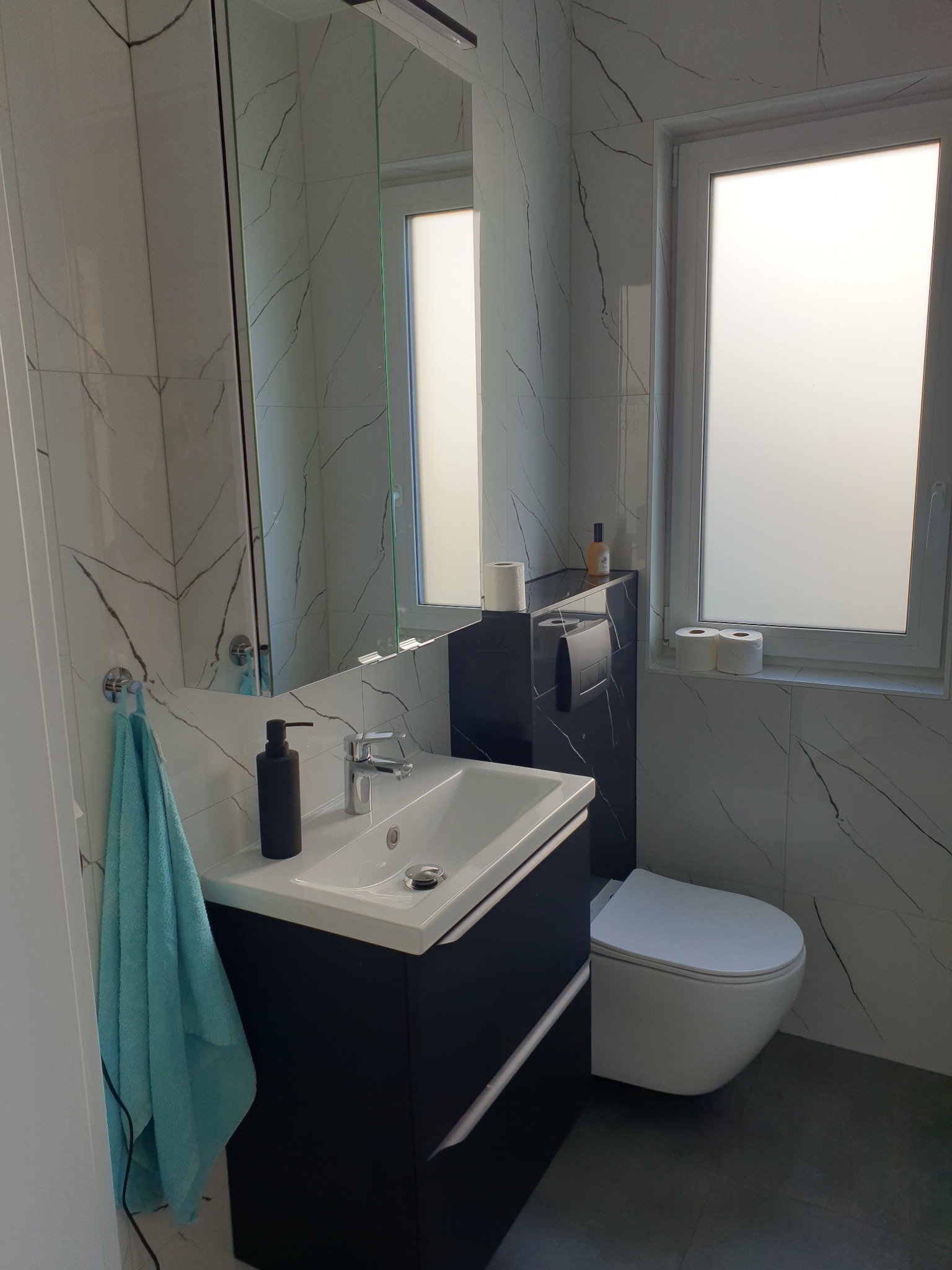 Nowoczesna łazienka z umywalką na szafce w kolorze granatowym, toaletą w zabudowie i lustrem. Ściany wyłożone białymi płytkami z szarym wzorem żyłkowania.