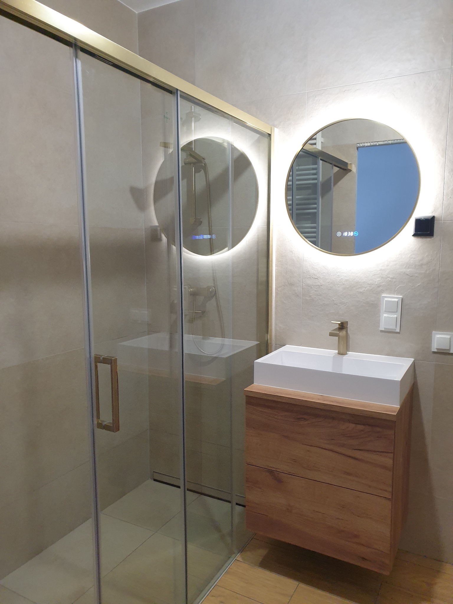 Nowoczesna łazienka z kabiną prysznicową ze złotymi ramami, okrągłym lustrem z podświetleniem LED i drewnianą szafką pod umywalką. Beżowe płytki na ścianach i podłodze.