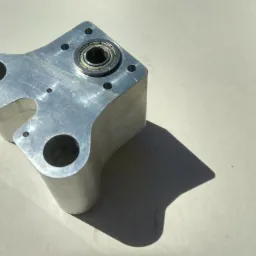 Aluminiowy element z łożyskiem, widoczne ślady obróbki i otwory montażowe, leżący na jasnym tle w ostrym świetle słonecznym.