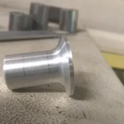 Metalowy element o kształcie tulei z kołnierzem, widoczne ślady obróbki, leżący na szorstkiej powierzchni, w tle inne metalowe detale.