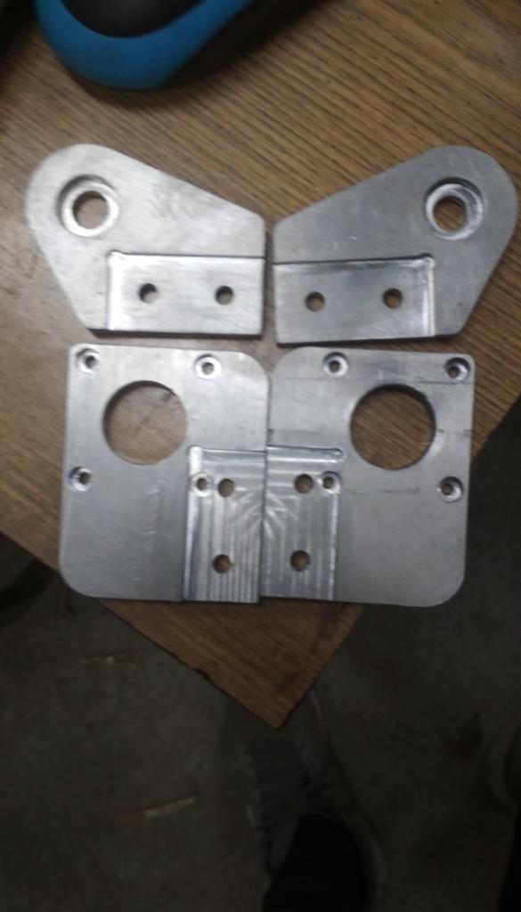 Cztery metalowe elementy z otworami, prawdopodobnie wykonane na zamówienie, leżące na drewnianej powierzchni.