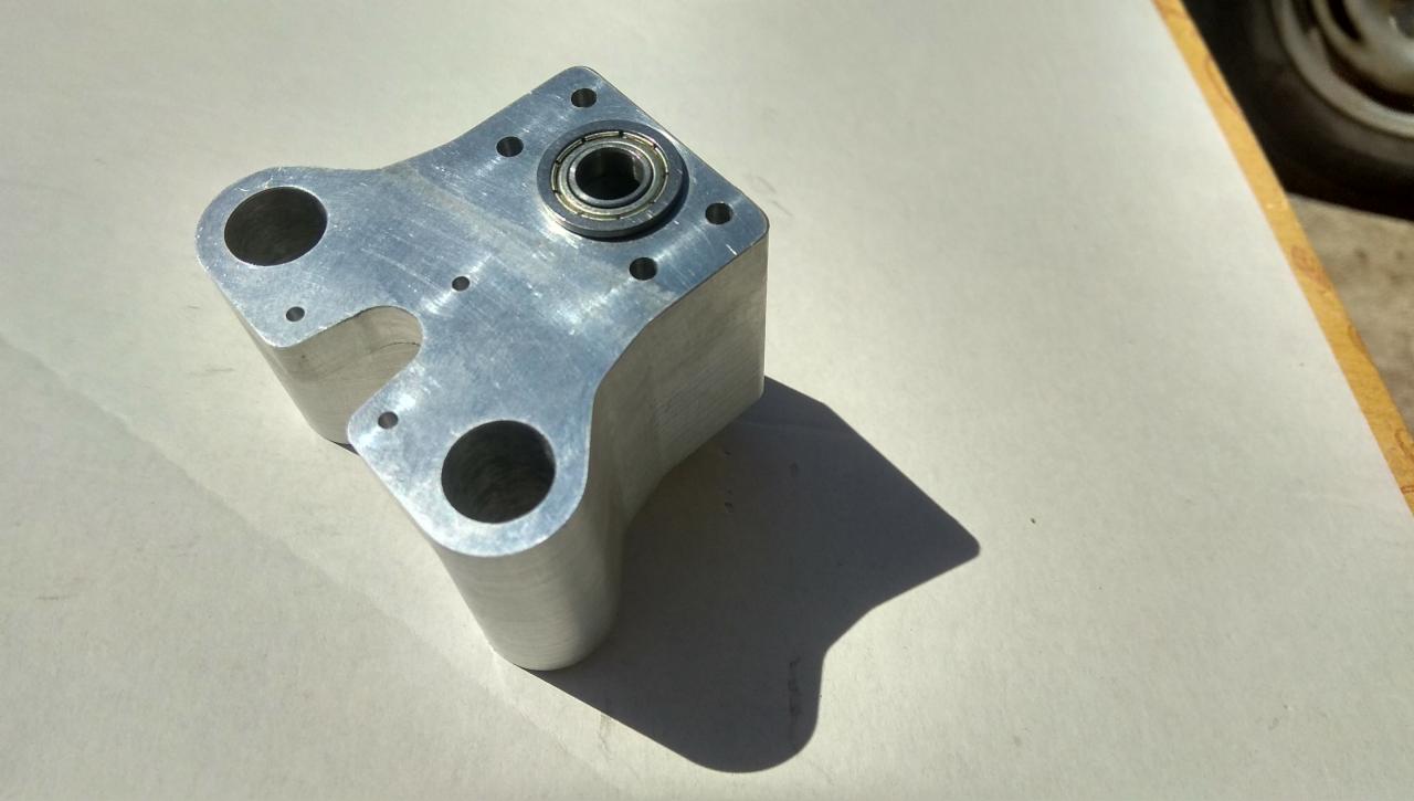 Aluminiowy element z łożyskiem, widoczne ślady obróbki i otwory montażowe, leżący na jasnym tle w ostrym świetle słonecznym.