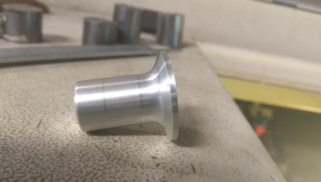 Metalowy element o kształcie tulei z kołnierzem, widoczne ślady obróbki, leżący na szorstkiej powierzchni, w tle inne metalowe detale.