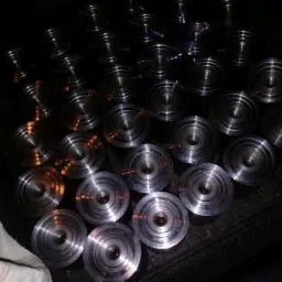 Partia obrobionych, metalowych, okrągłych elementów z centralnym otworem, ułożonych na czarnej, teksturowanej powierzchni. Widoczne ślady obróbki skrawaniem i odbicia światła.