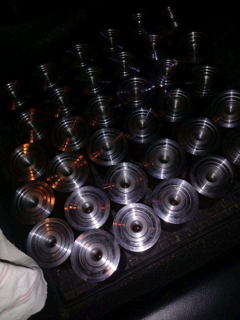 Partia obrobionych, metalowych, okrągłych elementów z centralnym otworem, ułożonych na czarnej, teksturowanej powierzchni. Widoczne ślady obróbki skrawaniem i odbicia światła.