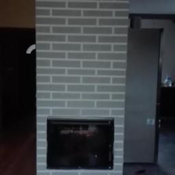 Fil_Bud - Pomalowany na szaro komin z imitacją cegły z czarnym wkładem kominkowym, na podłodze leżąca czarna listwa, w tle widoczne drzwi.