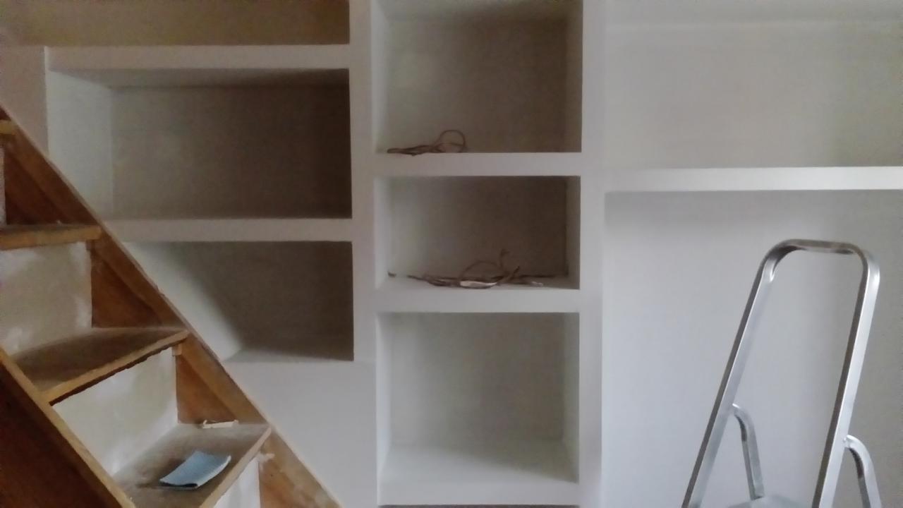 Białe, wnękowe półki pod schodami w trakcie wykańczania, z widoczną drewnianą konstrukcją schodów, aluminiową drabinką i drobnymi kablami wewnątrz półek.