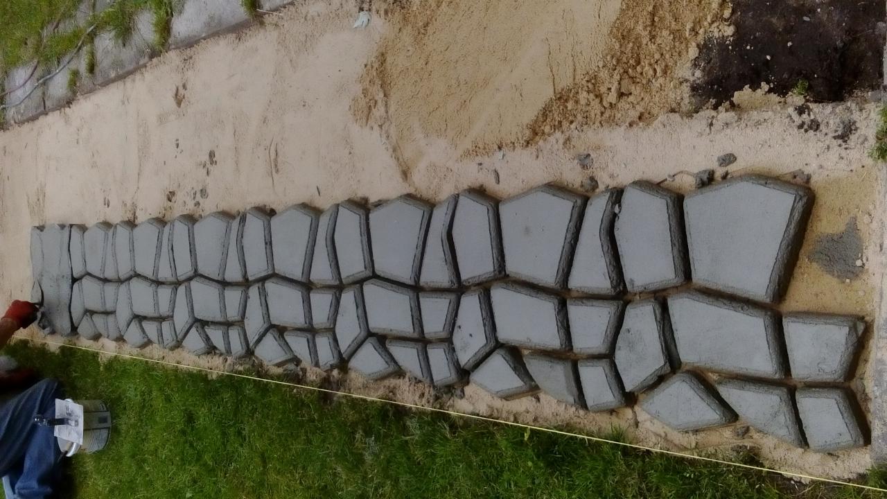 Układanie betonowych płyt chodnikowych o nieregularnych kształtach, widoczne ślady świeżego betonu, trawa i piasek w tle. Prace w toku.