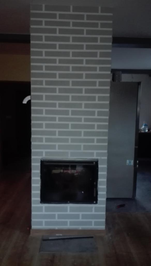 Pomalowany na szaro komin z imitacją cegły z czarnym wkładem kominkowym, na podłodze leżąca czarna listwa, w tle widoczne drzwi.
