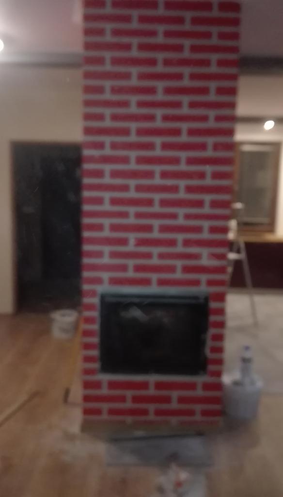 Fragment ściany z czerwonej cegły z otworem na kominek, widoczne ślady po remoncie i akcesoria malarskie w tle.