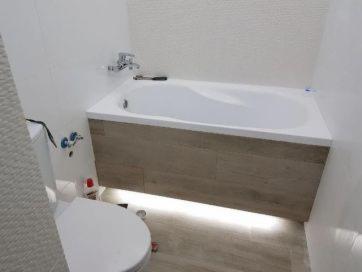 Biała wanna zabudowana panelami imitującymi drewno, z podświetleniem LED od dołu, w tle białe, teksturowane płytki ścienne. Widoczny fragment toalety i baterii wannowej.