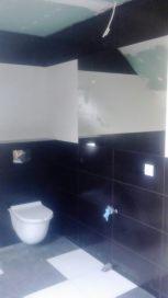 Częściowo wykończona łazienka z czarnymi płytkami ściennymi i białymi płytkami podłogowymi ułożonymi we wzór. Widoczne podwieszane WC, fragment sufitu z płyt gipsowo-kartonowych oraz niezakończone...