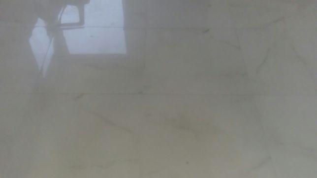 Błyszcząca, jasna podłoga z płytek imitujących marmur, odbijająca światło i fragment mebla w tle.