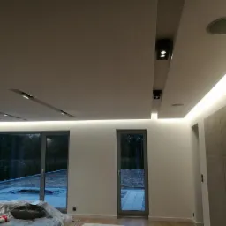 Nowoczesne wnętrze z podwieszanym sufitem, oświetleniem LED na obrzeżach i reflektorami punktowymi w czarnych obudowach. Widoczne duże okna z widokiem na zewnątrz.