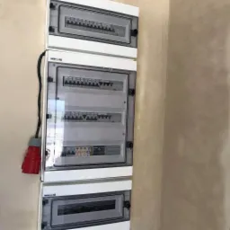 Trzy białe skrzynki rozdzielcze z przezroczystymi drzwiczkami zamontowane na beżowej ścianie, widoczny czerwony wtyk z kablem po lewej stronie.