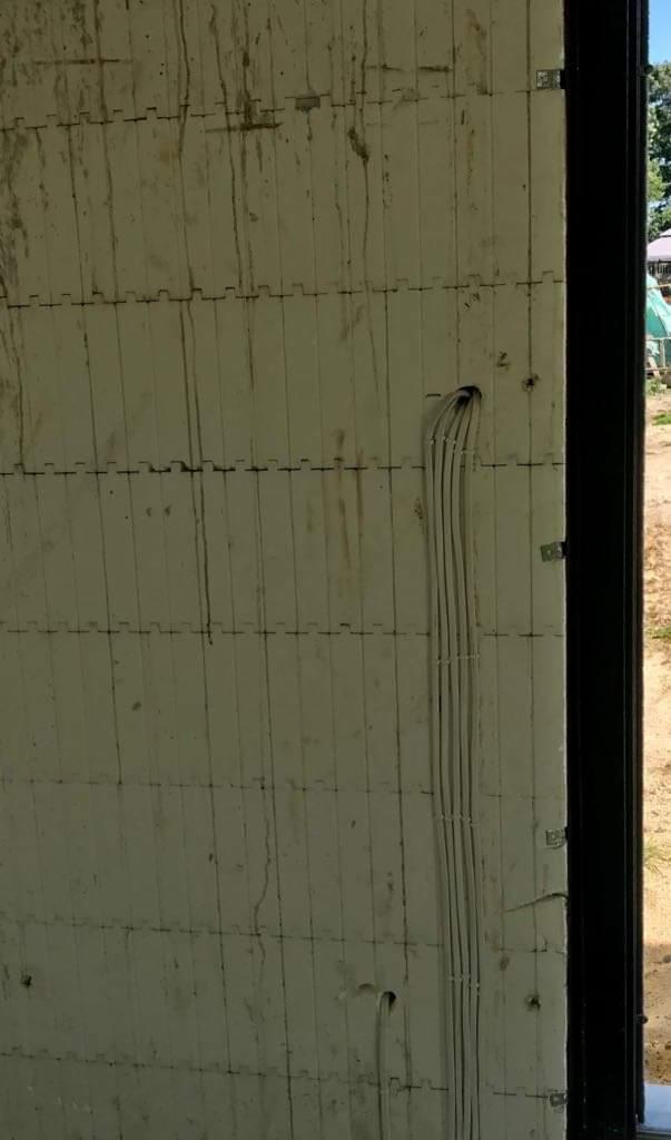 Biała ściana z widocznymi kablami elektrycznymi wychodzącymi z otworu, w tle widoczne elementy otoczenia zewnętrznego.