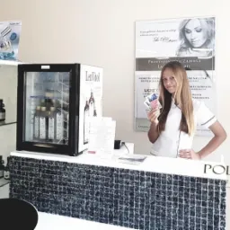 Recepcja gabinetu kosmetycznego z młodą recepcjonistką trzymającą karty lojalnościowe, za nią plakaty reklamowe zabiegów laserowych i chłodziarka z napojami, po bokach witryny z kosmetykami...