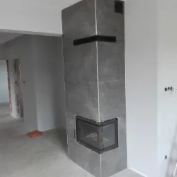 Obudowa kominka wykończona szarymi płytkami w surowym wnętrzu, widoczne niedokończone ściany i kabel na podłodze.