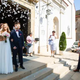 Para młoda wychodzi z kościoła, obsypywana konfetti w kształcie motyli, goście weselni w tle, mężczyzna odpala tubę z konfetti.