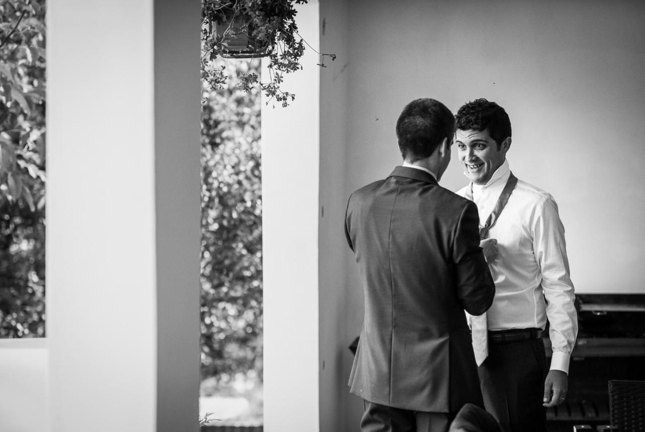 Czarno-białe zdjęcie mężczyzny w białej koszuli, poprawiającego krawat, z uśmiechem na twarzy, podczas gdy drugi mężczyzna w garniturze stoi przed nim, prawdopodobnie pomagając mu się przygotować.