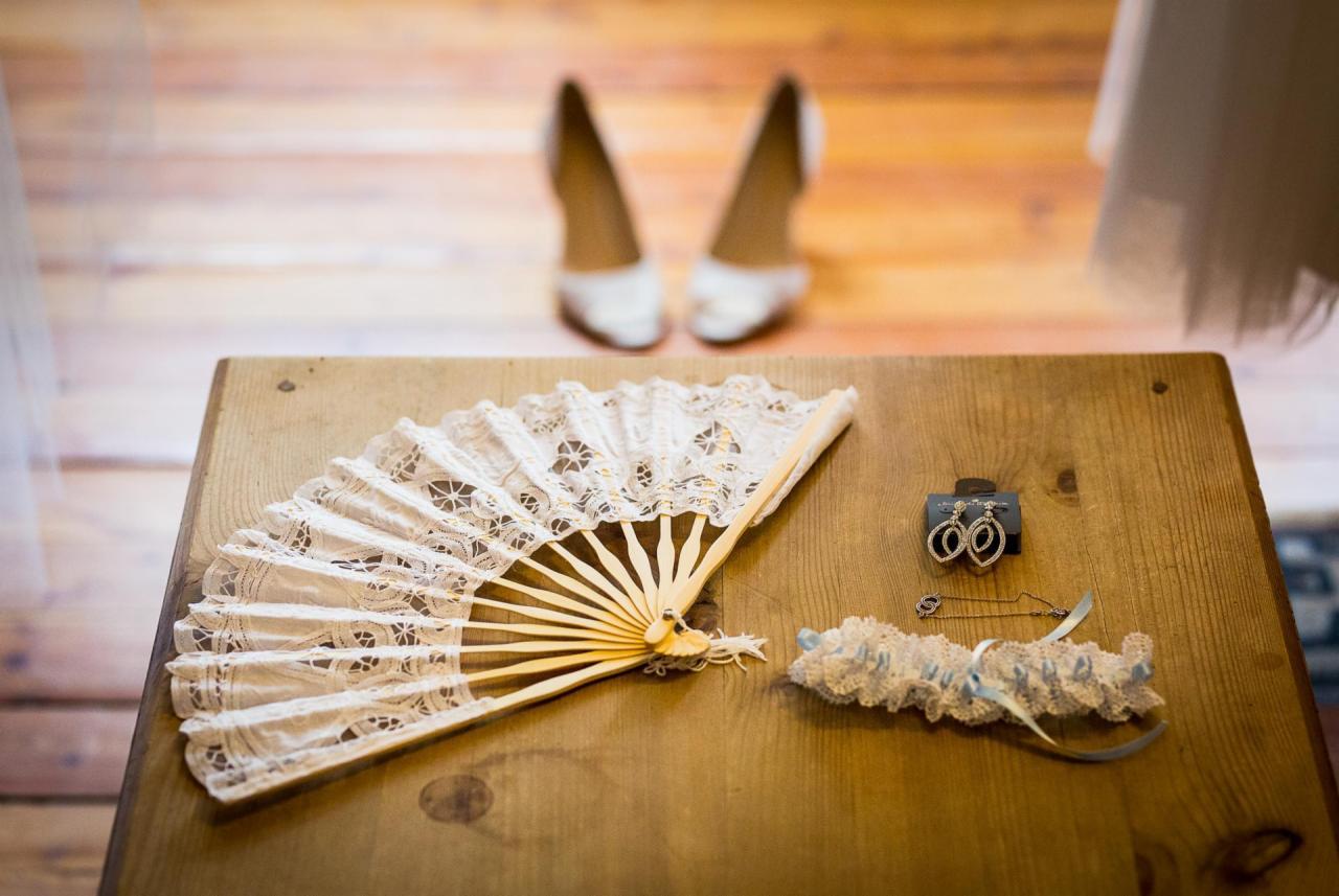 Przygotowania ślubne: biały wachlarz z koronką, kolczyki w pudełku, podwiązka i szpilka na drewnianym stole, w tle białe szpilki i fragment sukni.
