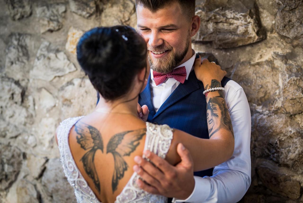 Uśmiechnięty pan młody w granatowej kamizelce i bordowej muszce przytula pannę młodą z tatuażem skrzydeł na plecach, na tle kamiennej ściany.