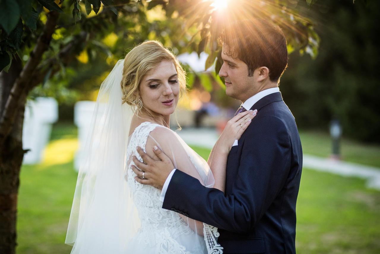 Panna młoda w koronkowej sukni i welonie obejmuje pana młodego w granatowym garniturze, promienie słońca przebijają przez liście drzewa.