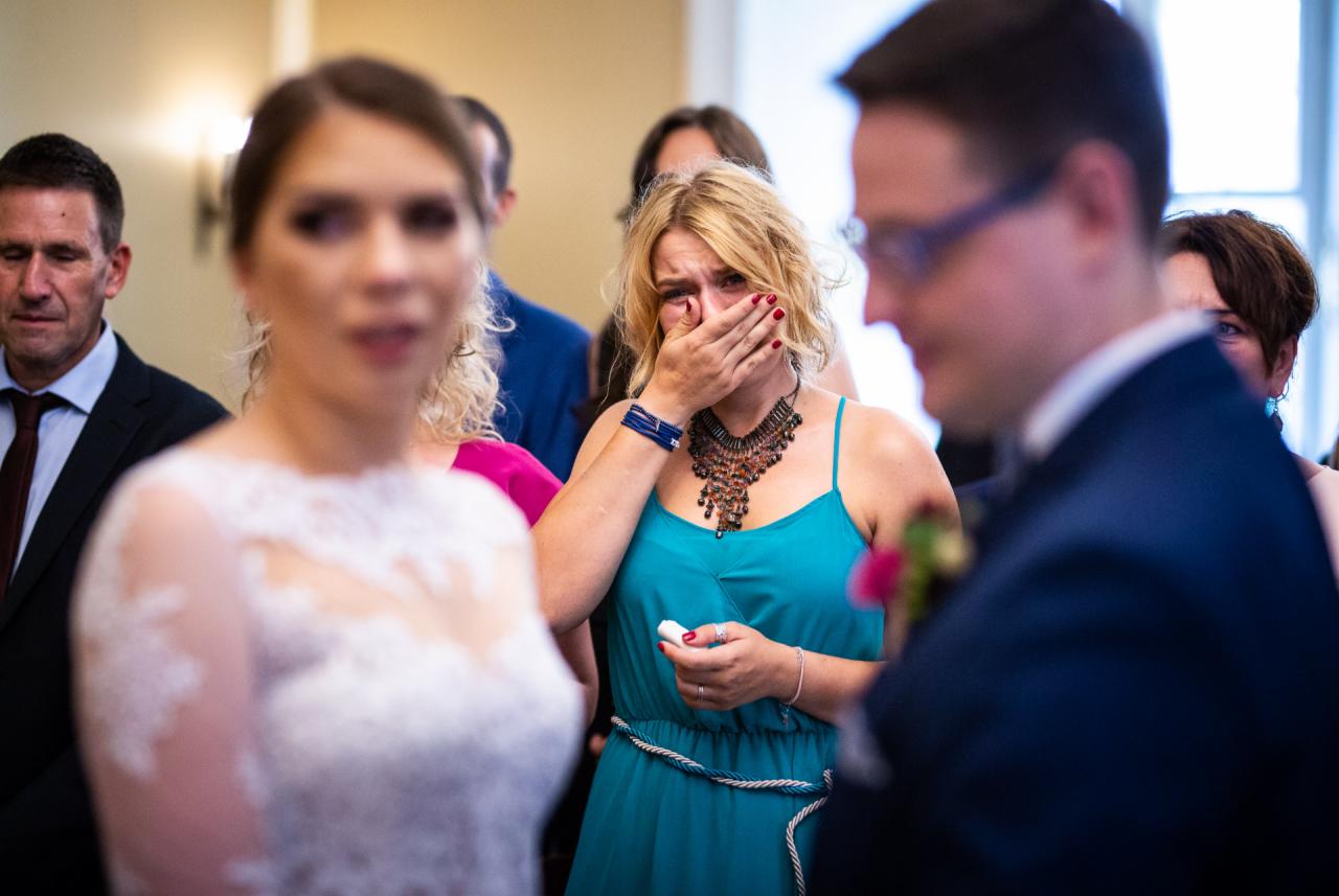 Wzruszona kobieta w turkusowej sukience, zakrywająca twarz dłonią podczas ceremonii ślubnej, w tle rozmazana panna młoda w białej sukni i goście.
