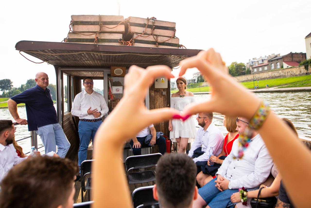 Goście na barce tworzą serce dłońmi, w tle panna młoda w białej sukience i mężczyźni w koszulach, widok na rzekę i budynki.