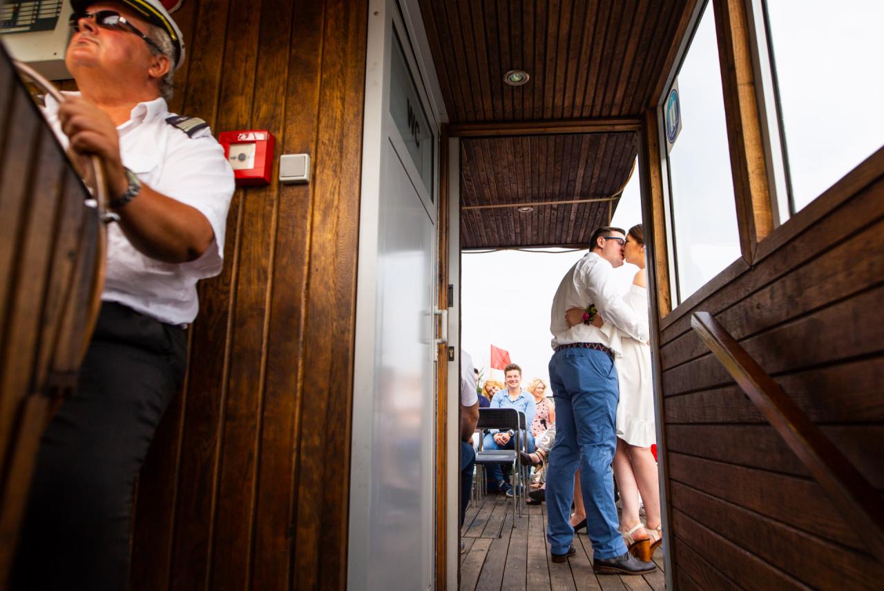 Para młoda całuje się na statku podczas ceremonii ślubnej, kapitan statku stoi przy sterze, w tle goście.