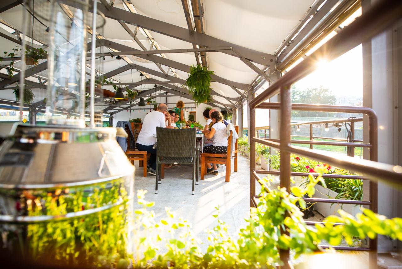 Grupa osób siedzi przy stole na zewnątrz, pod zadaszeniem z metalową konstrukcją i wiszącymi roślinami. W tle widoczny zachód słońca i roślinność.