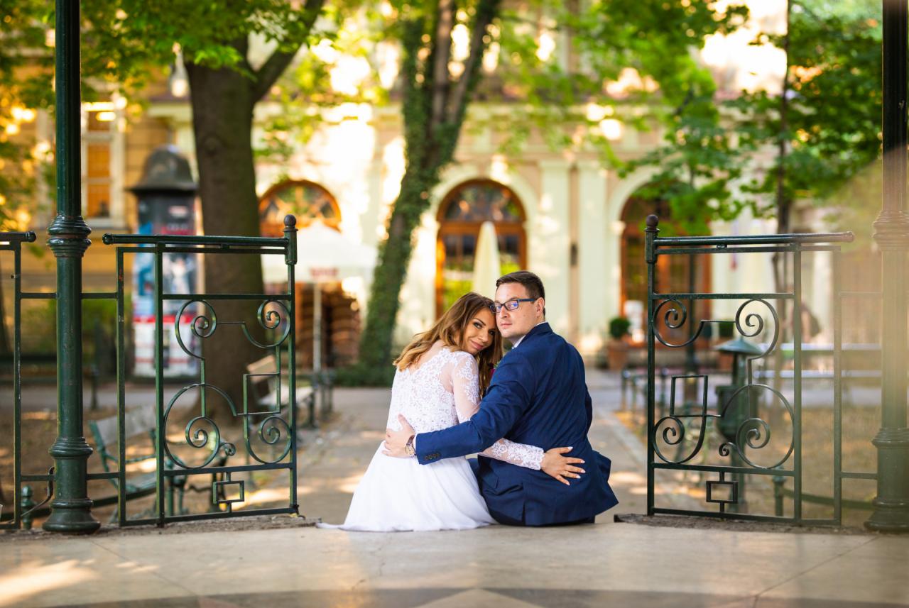 Para młoda siedzi na chodniku, obejmując się na tle ozdobnej, niskiej metalowej barierki i budynku w parku.