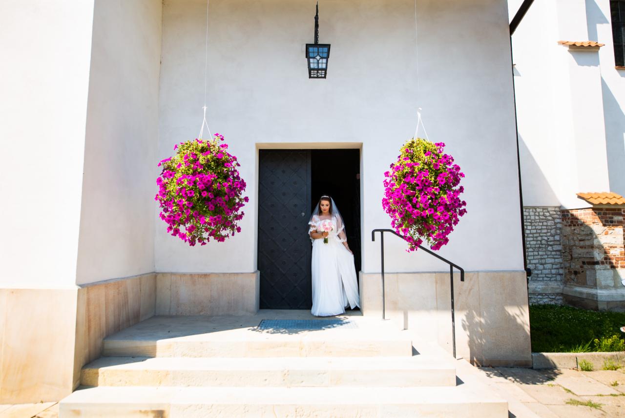 Panna młoda w białej sukni wychodzi z kościoła, ozdobionego wiszącymi koszami z różowymi kwiatami, trzymając bukiet.
