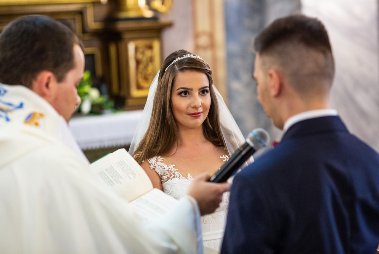 Panna młoda w białej sukni i welonie, z tiarą we włosach, podczas ceremonii ślubnej w kościele, spogląda na pana młodego trzymającego mikrofon, obok ksiądz z księgą.