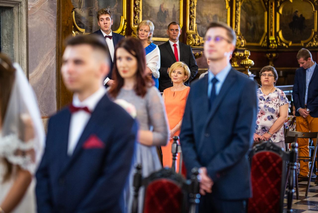 Goście weselni w kościele, mężczyźni w garniturach i muchach, kobiety w sukienkach, widoczna część welonu panny młodej.