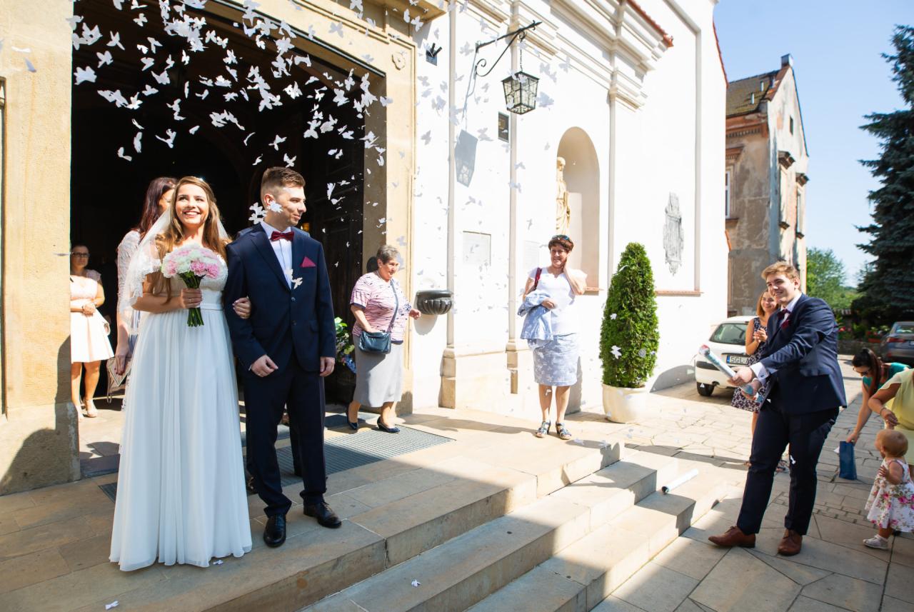 Para młoda wychodzi z kościoła, obsypywana konfetti w kształcie motyli, goście weselni w tle, mężczyzna odpala tubę z konfetti.