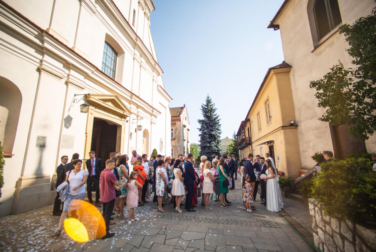 Goście weselni zgromadzeni przed kościołem po ceremonii, obsypani konfetti, panna młoda trzyma bukiet.