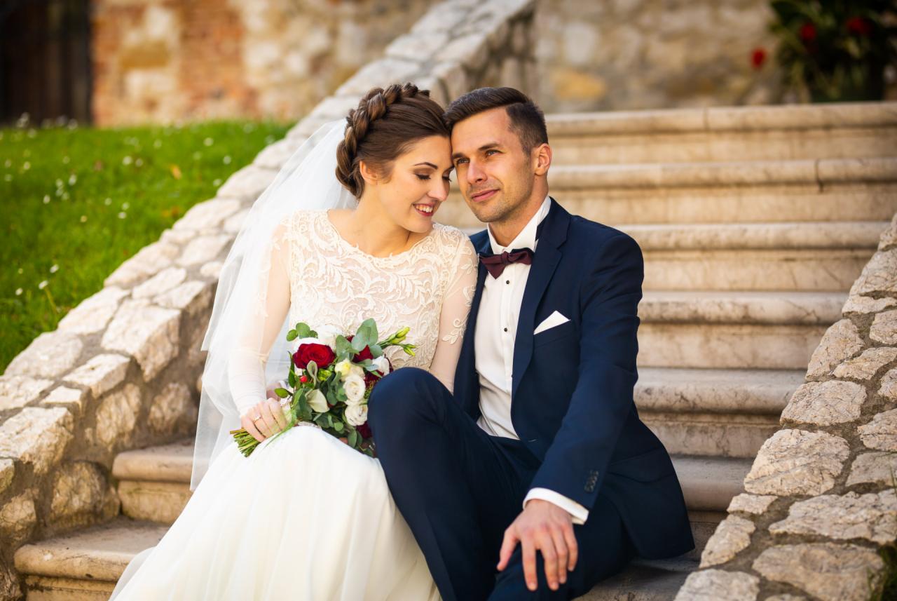 Para młoda siedzi na kamiennych schodach; panna młoda w koronkowej sukni trzyma bukiet czerwonych i białych róż, pan młody w granatowym garniturze z bordową muchą.