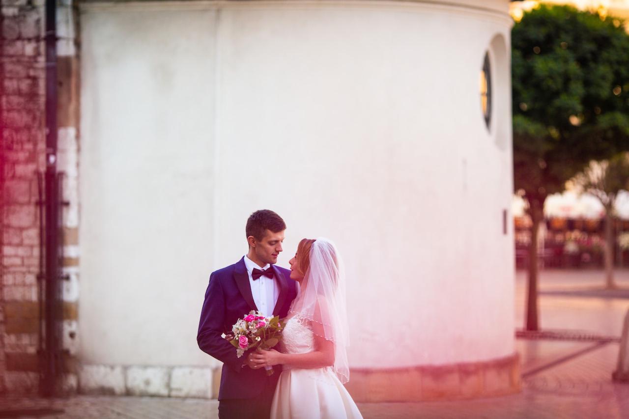 Para młoda wpatrzona w siebie, on w granatowym smokingu z bukietem, ona w sukni ślubnej z welonem, na tle białej, okrągłej budowli z małym oknem i drzewami w tle.